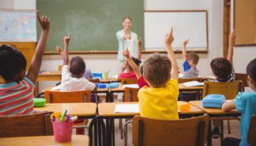 students with their hands raised in a classroom as the teacher at the front of the class points to someone